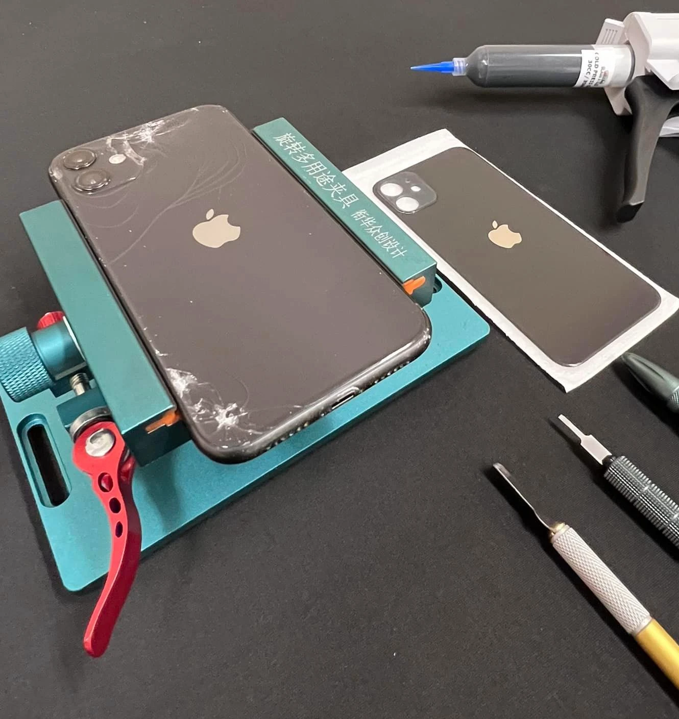 Cracked iPhone back glass and a replacement panel on a workbench